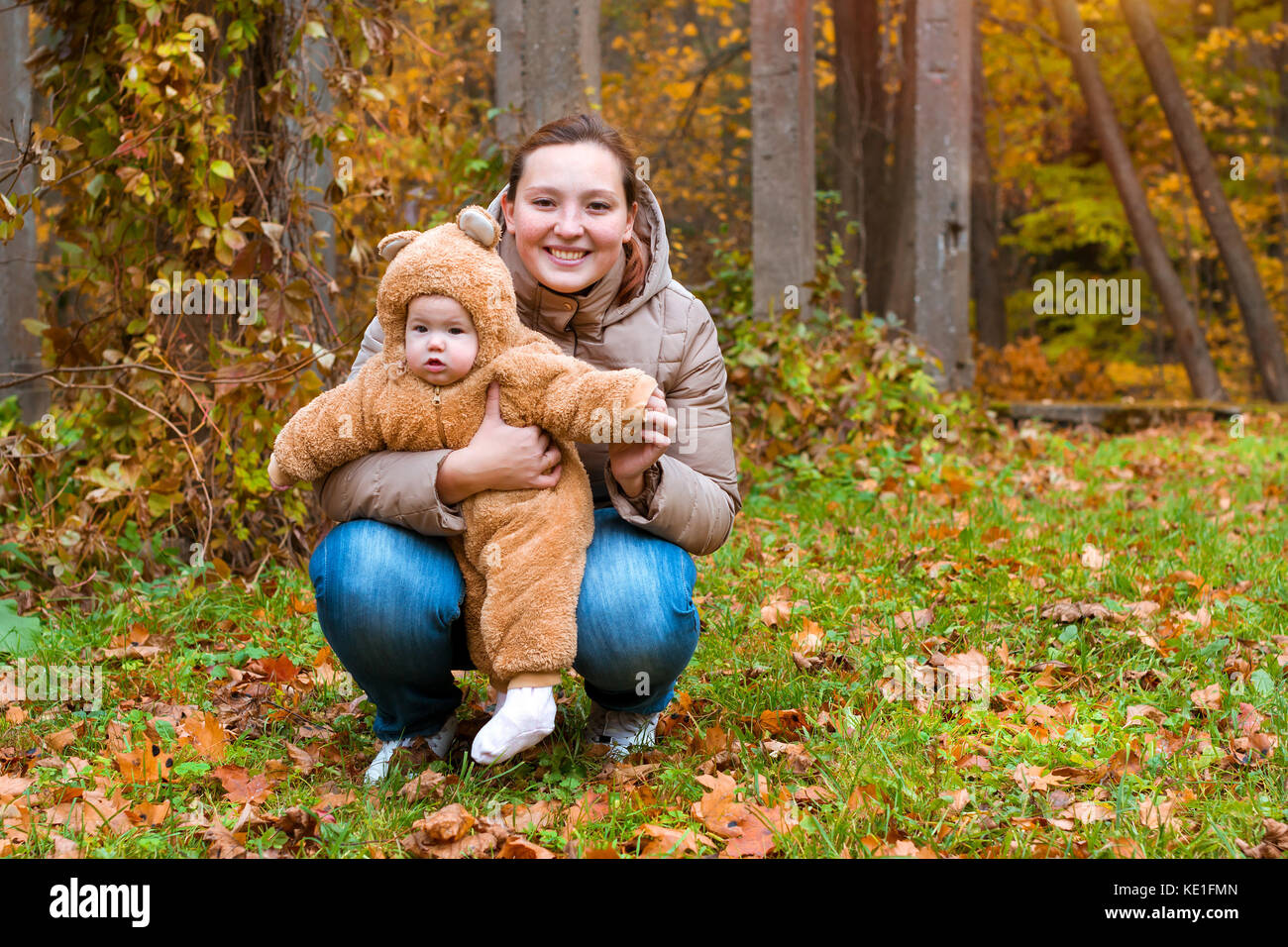 Donna con bambino in armi, ponendo sullo sfondo del parco d'autunno. bambino vestito in caldi stilizzata di Teddy bear costume, divertirsi sulla madre di braccia. famiglia w Foto Stock