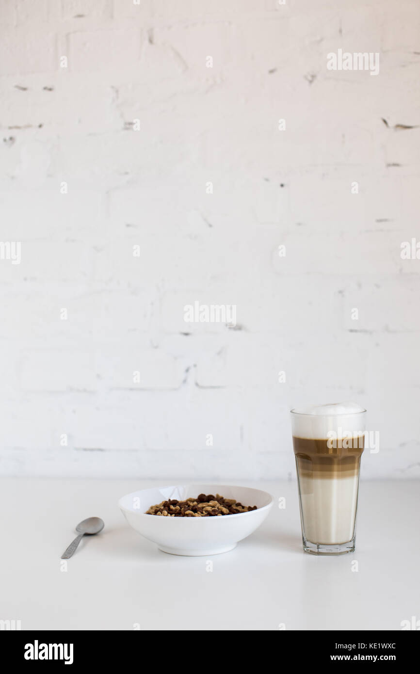 Bicchiere di caffè latte e cornflakes nella ciotola con il cucchiaio sul tavolo bianco Foto Stock