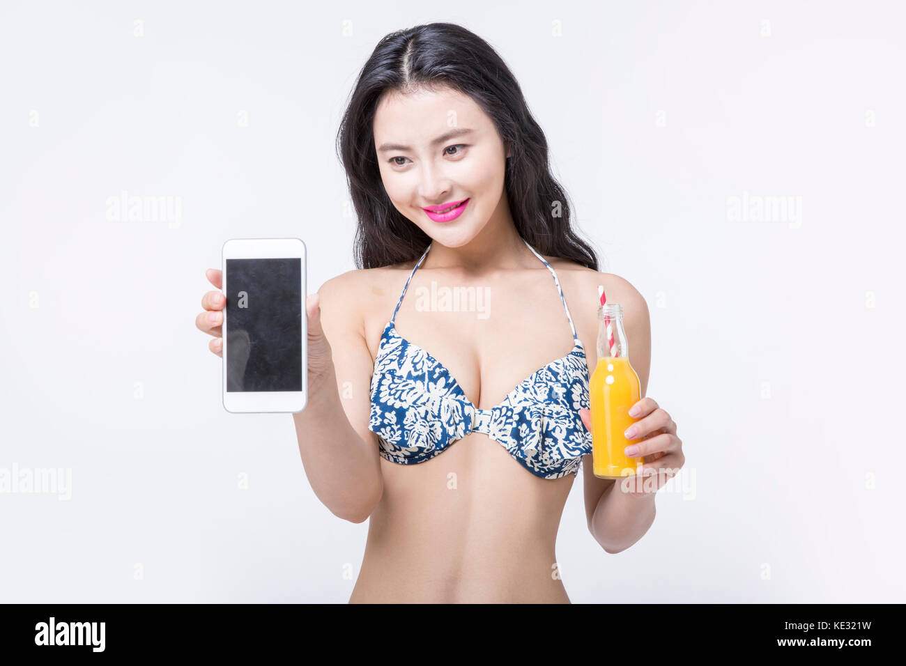 Sorridente giovane donna slim in bikini in posa con uno smartphone e succo di arancia Foto Stock