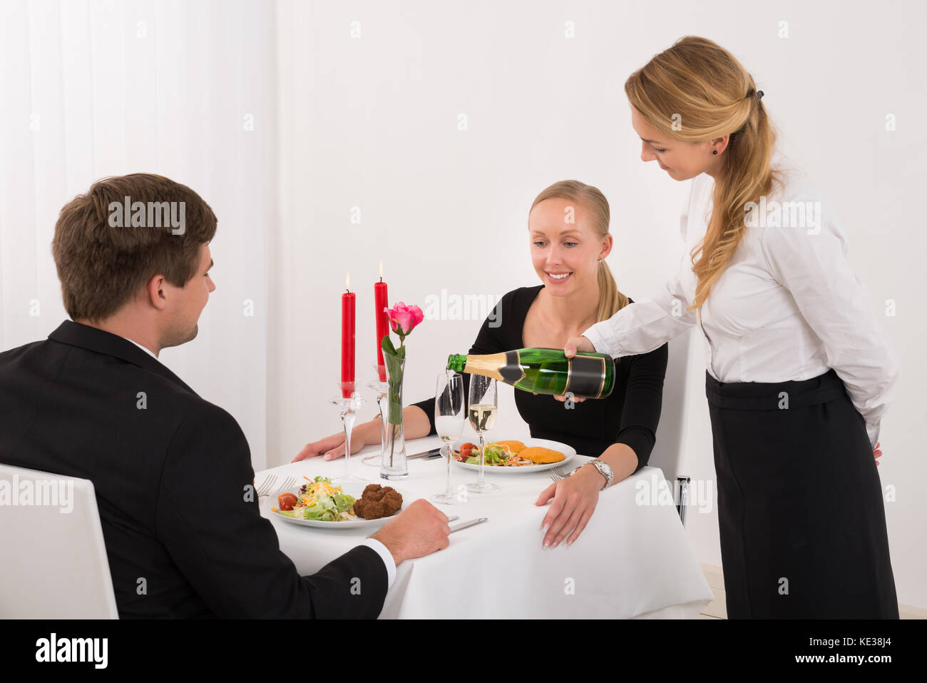 Cameriera femmina versando Champagne in vetro per la felice coppia giovane nel ristorante Foto Stock