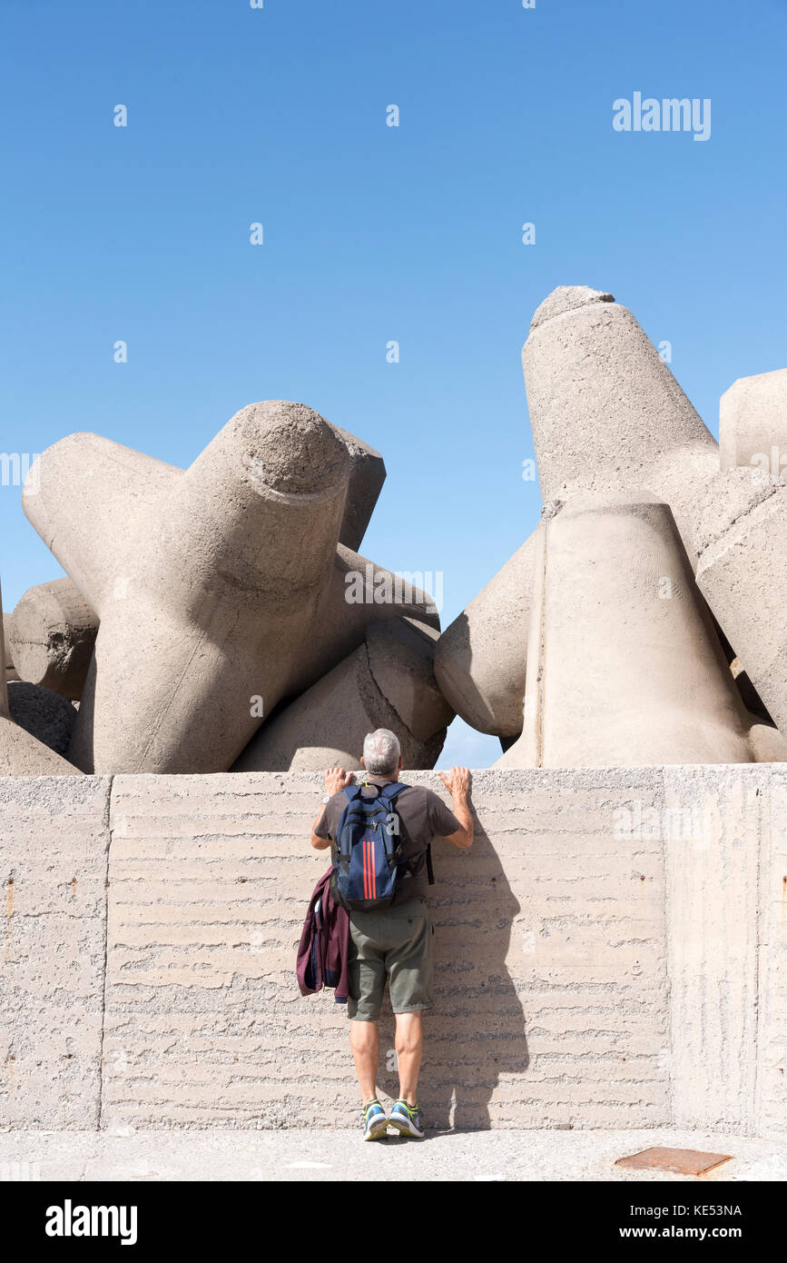 L'uomo sulle punte delle dita ispezionando l'uomo fatto blocchi di cemento, Tetrapods, che agiscono come difesa del mare intorno ad un muro del porto. Creta, Grecia, 2017 Foto Stock