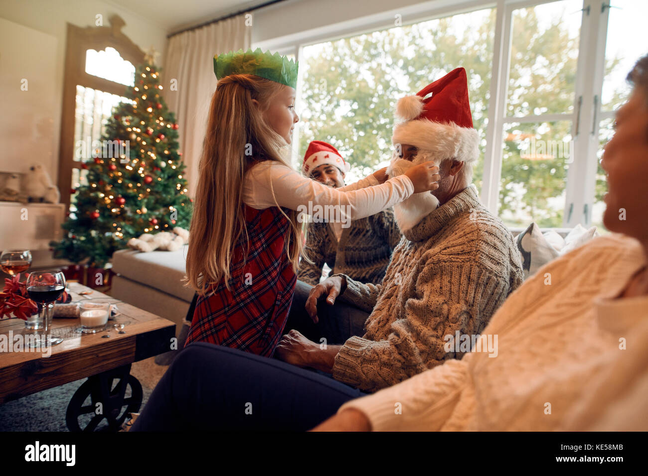 Bambina mettendo fake santa claus di barba e hat sul suo nonno. Famiglia insieme seduta nel salotto della vigilia di Natale. Foto Stock