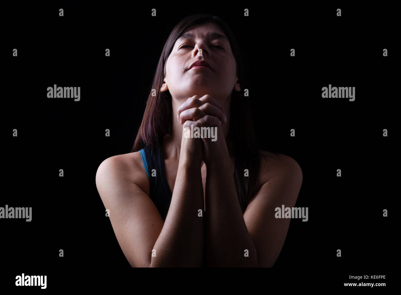 Close up di una fedele donna orante, mani giunte in preghiera, di testa in alto e gli occhi chiusi in fervore religioso, su uno sfondo nero. preghiera speranza pregare Foto Stock