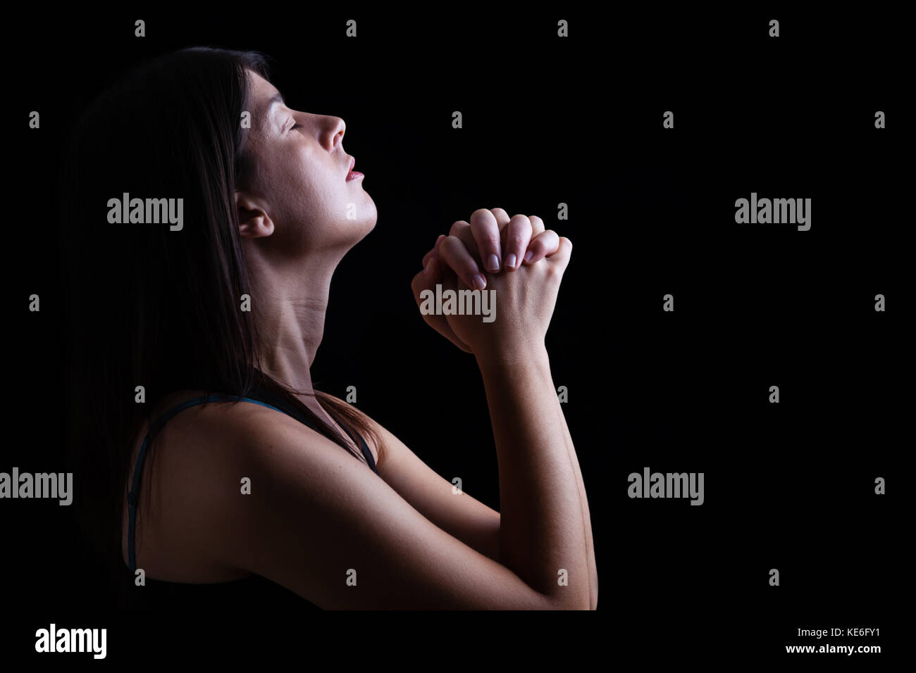 Close up di una fedele donna orante, mani giunte in preghiera, di testa in alto, gli occhi chiusi nel fervore religioso. Sfondo nero la preghiera cristiana e religiosa Foto Stock