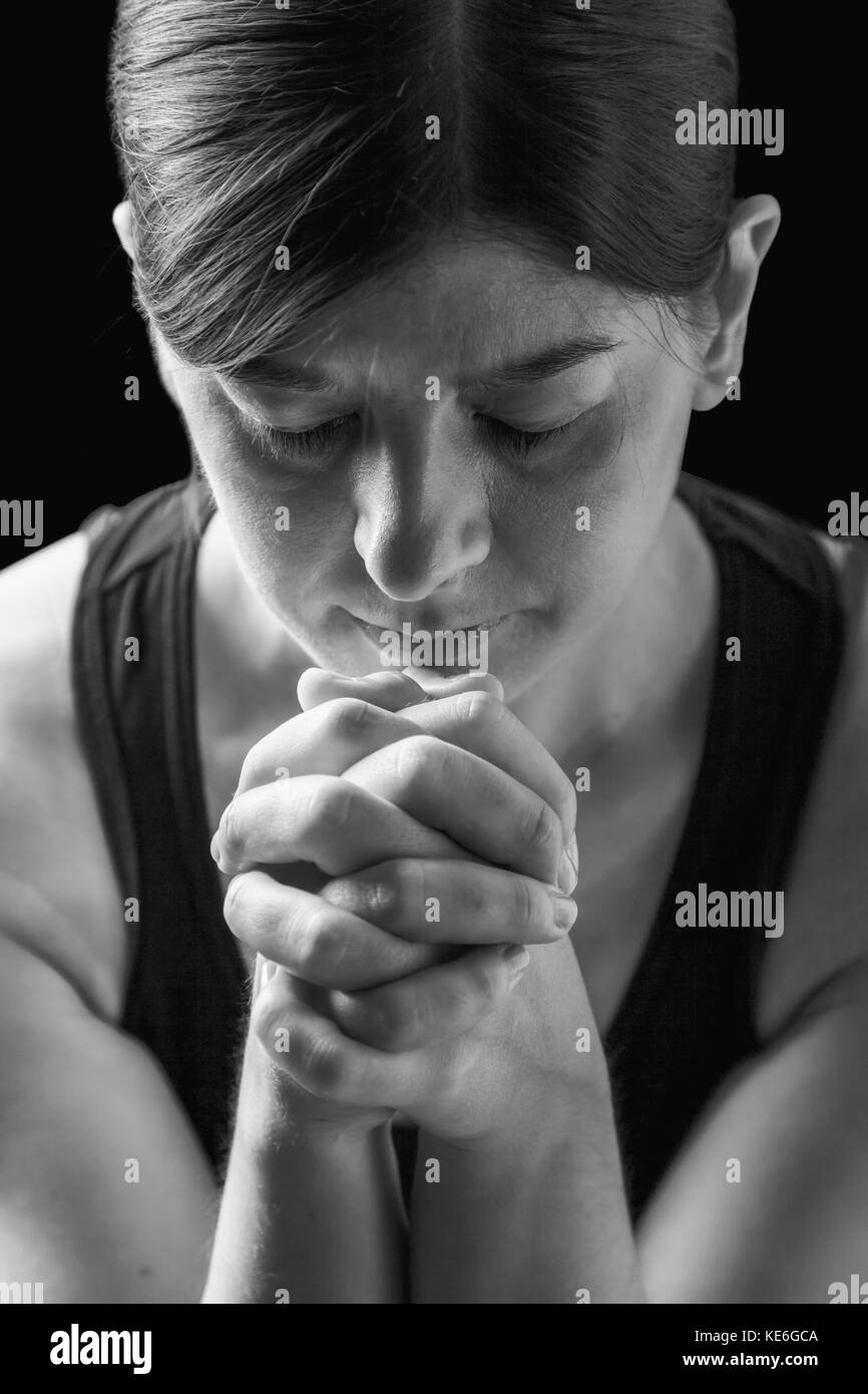 Tasto basso ritratto di una donna di fedeli di pregare, mani giunte nel culto a Dio, a testa in giù e gli occhi chiusi in fervore religioso, su uno sfondo nero. Foto Stock