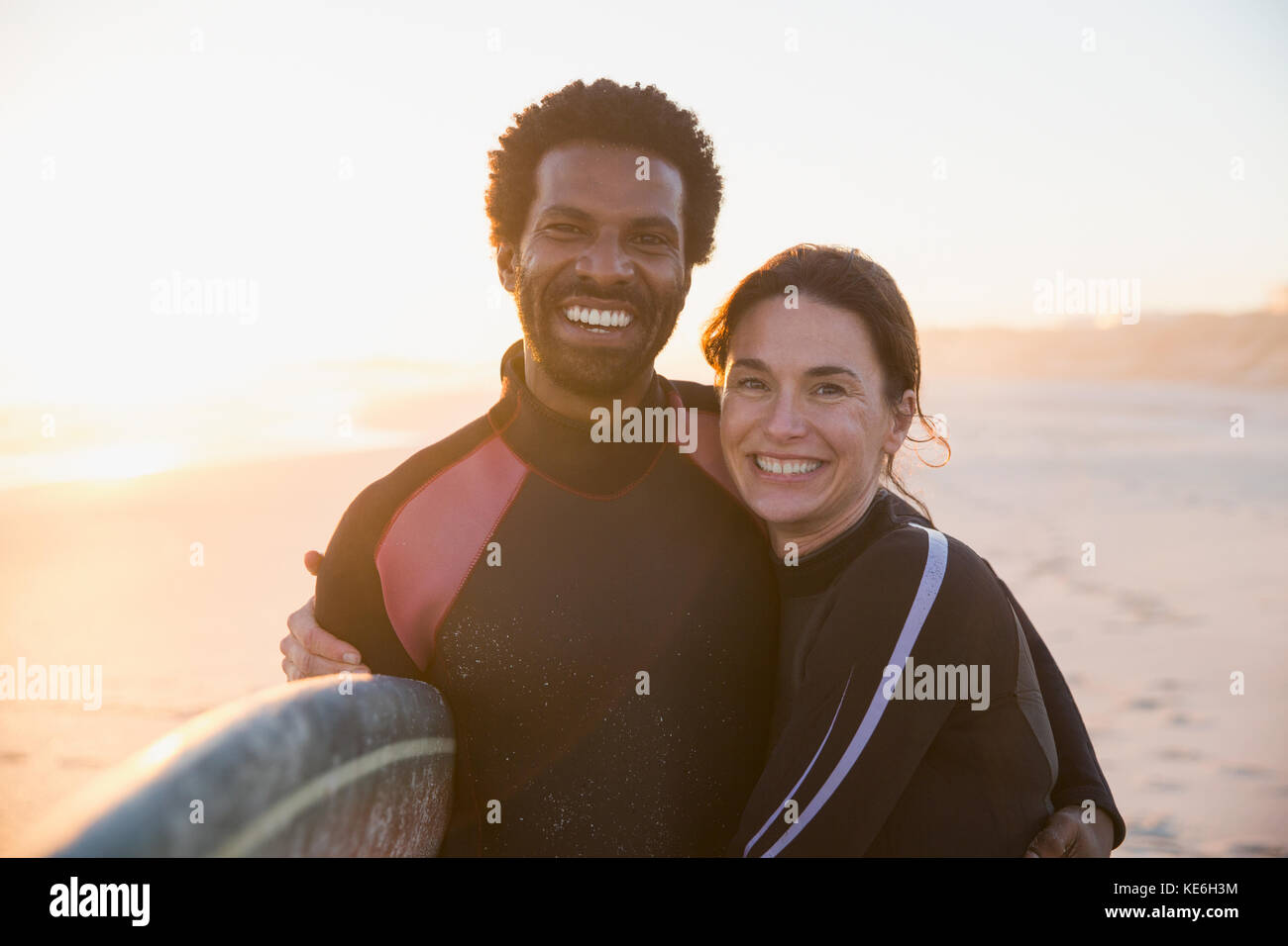 Ritratto sorridente, fidata coppia multietnica con tavola da surf sulla soleggiata spiaggia del tramonto estivo Foto Stock