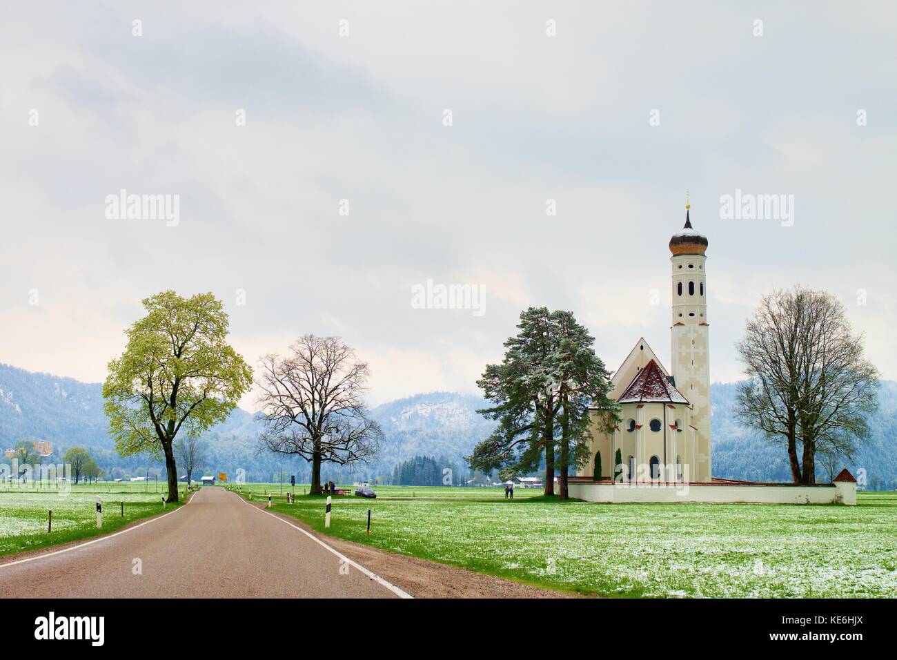 Vecchia chiesa e cimitero. dritta strada asfaltata a prati. aprile meteo. primavera umida di neve in già fresco verde erba montagne a soffietto. Foto Stock