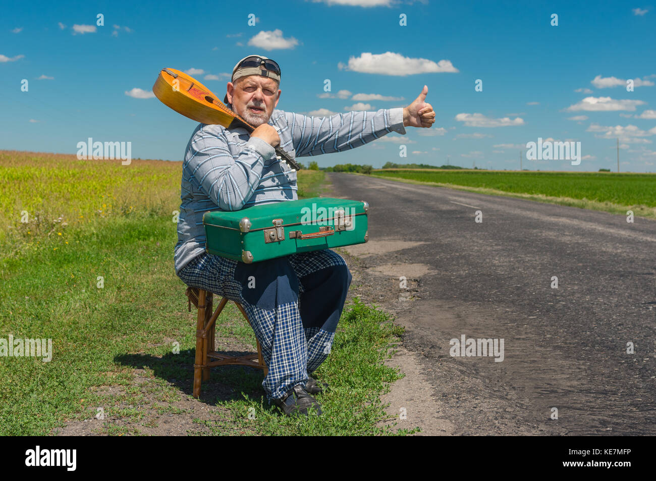 Positivo il sollevatore senior-escursionista avente breve riposo su una strada rurale con un antico valigia verde e il mandolino Foto Stock