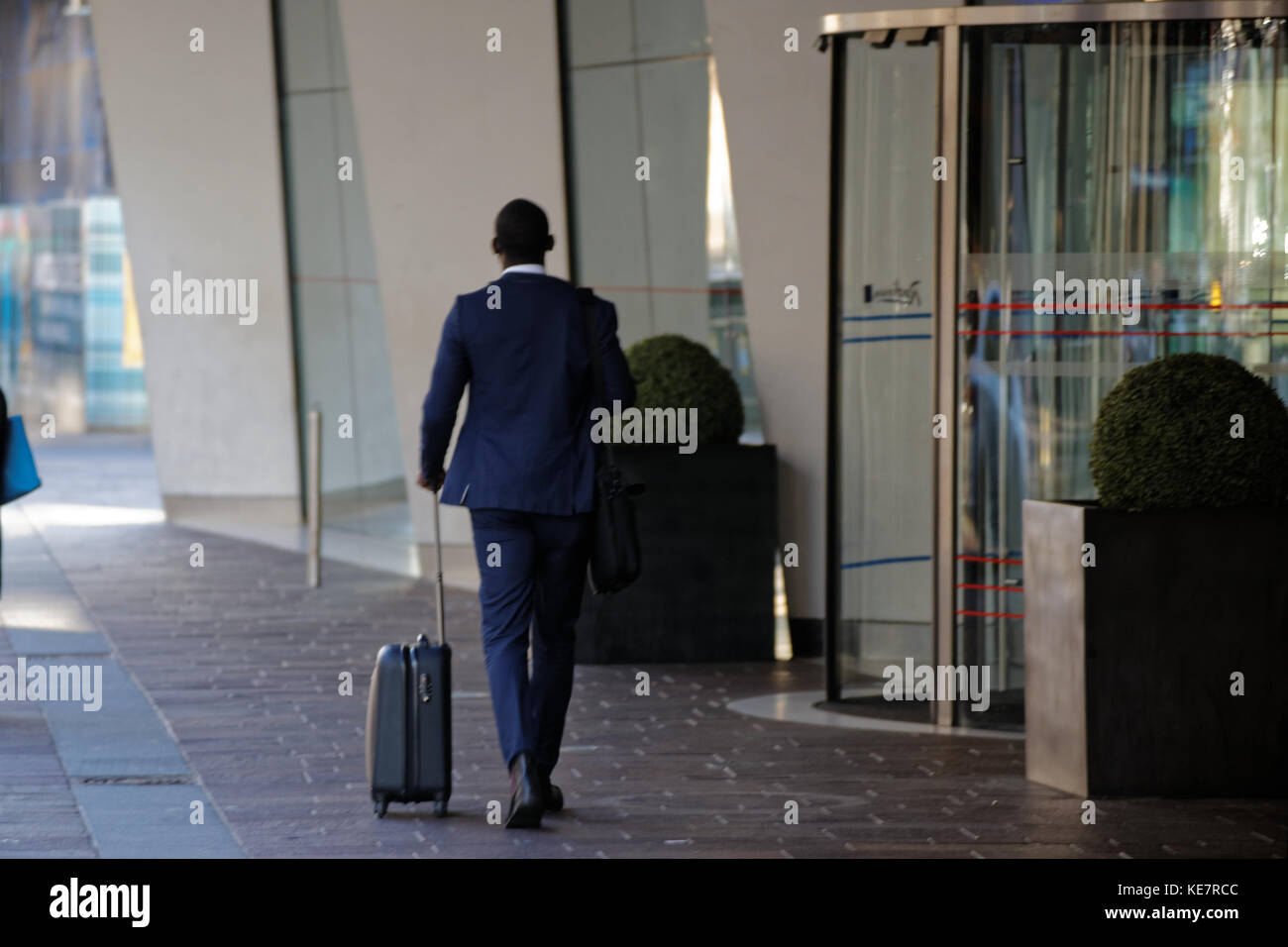Imprenditore nero in una tuta blu con trolley bag entrando in uscita hotel porta girevole entrata Radisson Blu Hotel, Glasgow Foto Stock