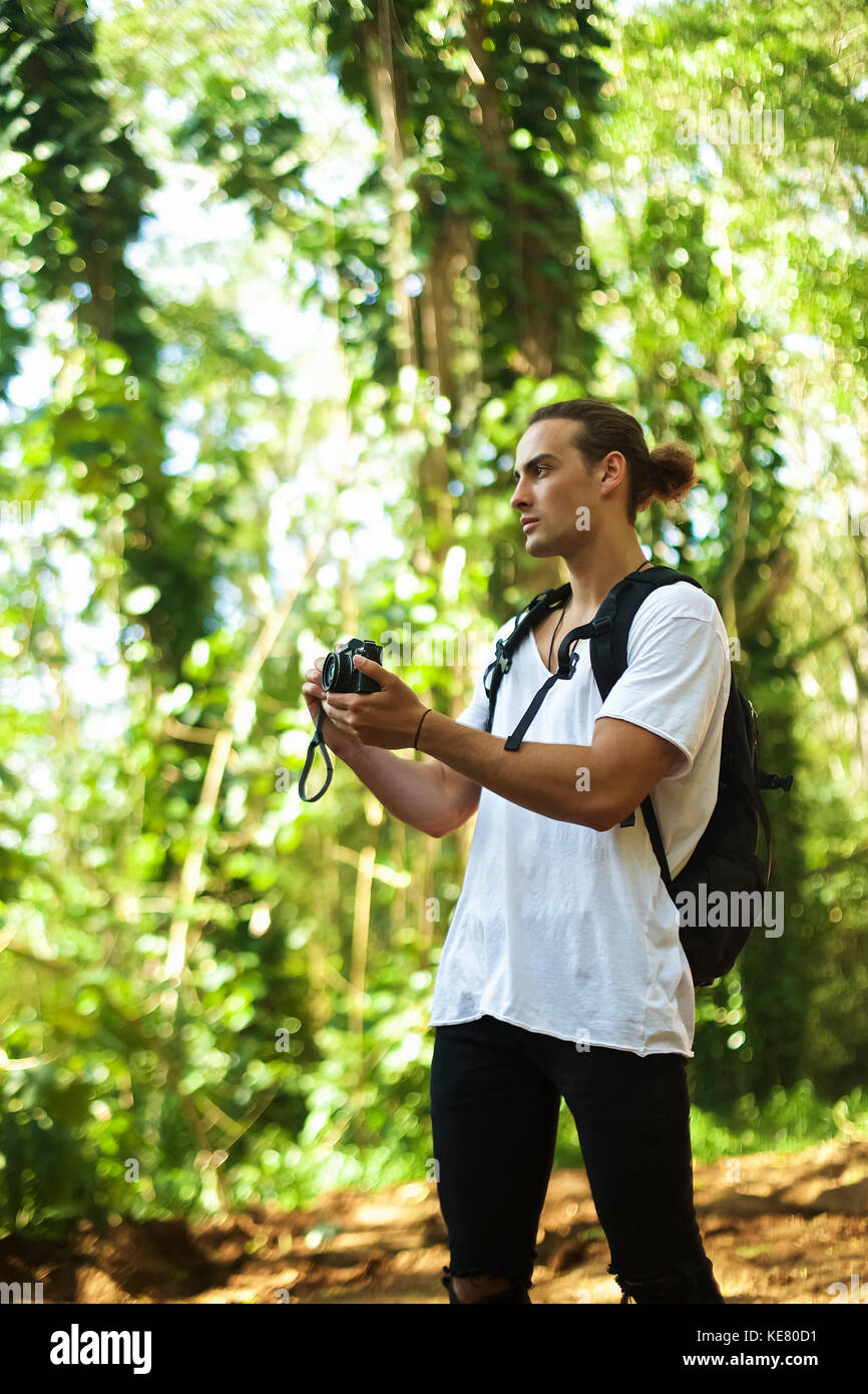 Un giovane uomo sorge su un sentiero che indossa uno zaino e scattare una foto con una fotocamera durante una escursione; Hawaii, Stati Uniti d'America Foto Stock