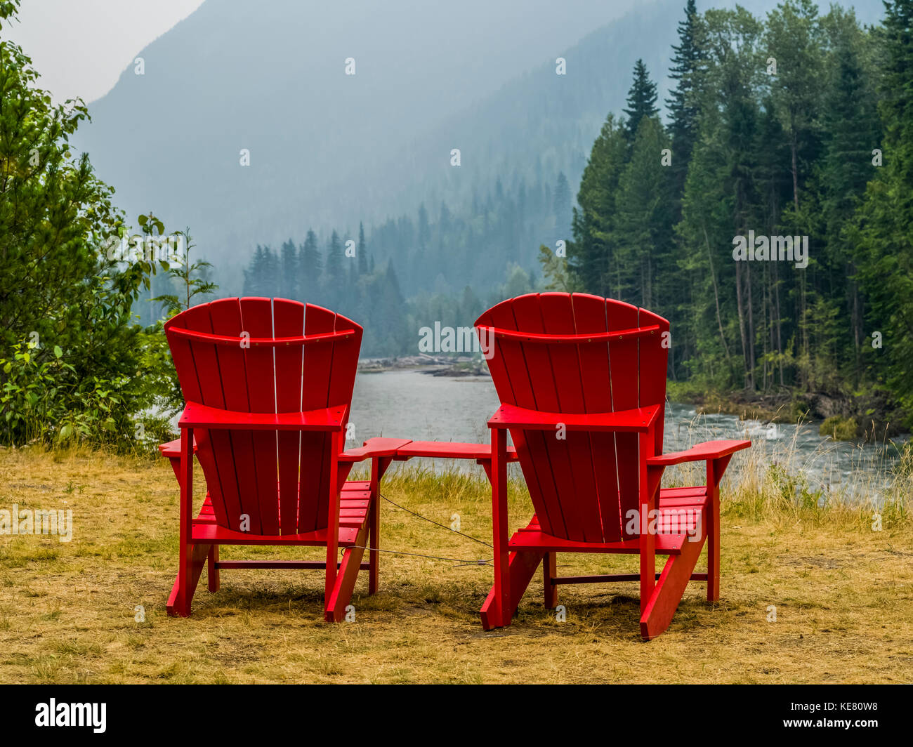 Due rossi sedie adirondack seduto sul prato che si affaccia sul fiume Columbia e coperta di foresta montagne; Revelstoke, British Columbia, Canada Foto Stock