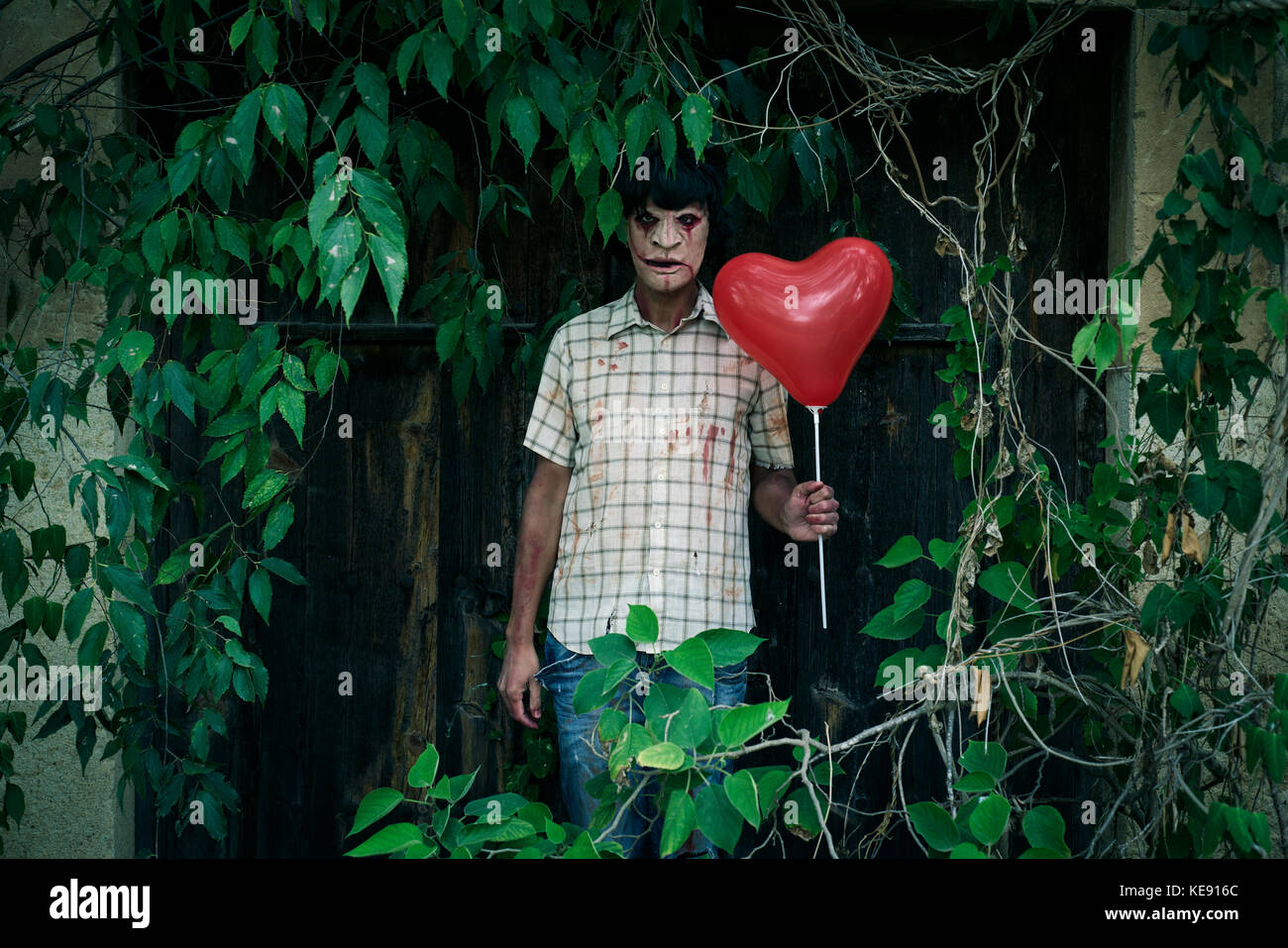 Un pauroso sfigurato uomo con un cuore rosso palloncino a forma di di fronte alla porta di una casa abbandonata coperti da erbacce Foto Stock