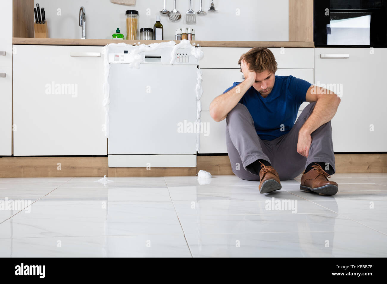 Giovane uomo sconvolto seduto accanto alla lavastoviglie con schiuma che esce in cucina Foto Stock