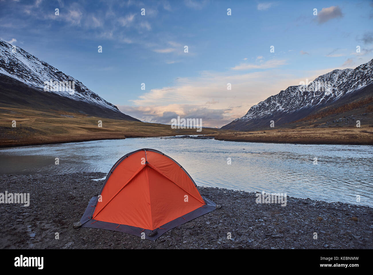 Campeggio e un arancio tenda vicino a un fiume con cime innevate in montagna Foto Stock