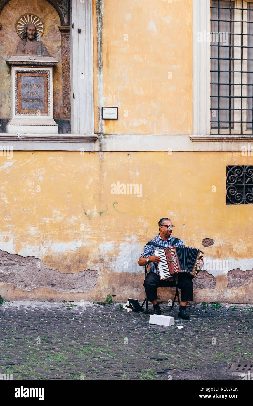 Un uomo suona la fisarmonica nel quartiere Ghetto di Roma, Italia. una statua di Gesù guarda verso il basso su di lui. Foto Stock