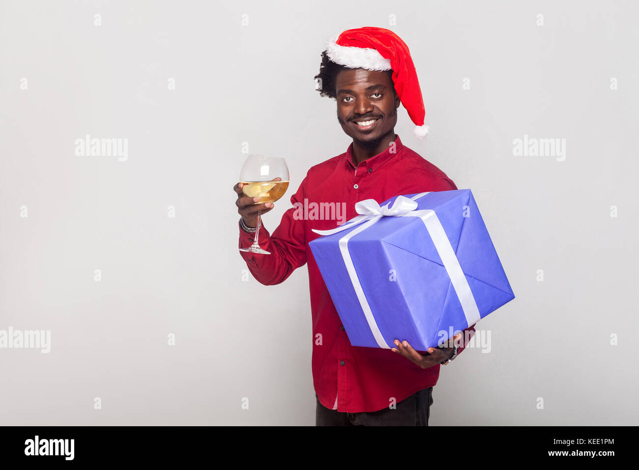 La felicità afro uomo in rosso di trattenimento tappo champagne in vetro e confezione regalo, guardando la fotocamera e toothy sorridente. studio shot. sfondo grigio Foto Stock