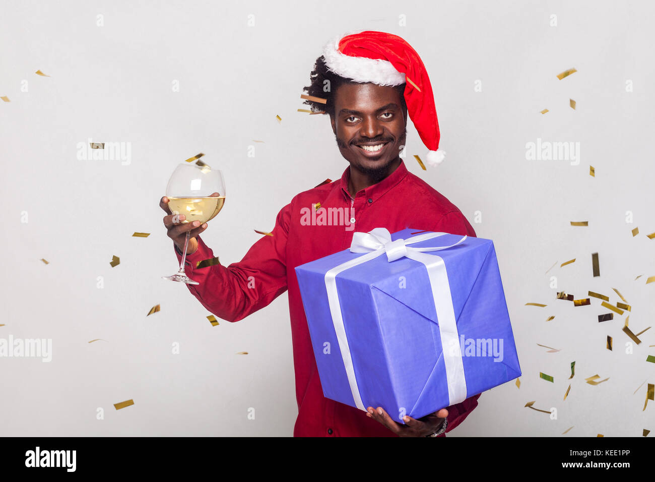 Intorno la felicità afro uomo nel cappuccio rosso, mosche metaphane oro, uomo con vetro di champagne e confezione regalo, guardando la fotocamera e toothy sorridente. studio sho Foto Stock