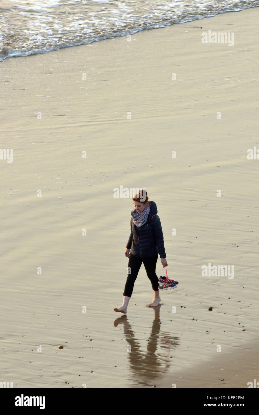 Una donna che cammina lungo una spiaggia sul suo proprio al tramonto sguazzare nel surf che porta le sue scarpe per evitare il loro contatto bagnato al mare in riva al mare Foto Stock