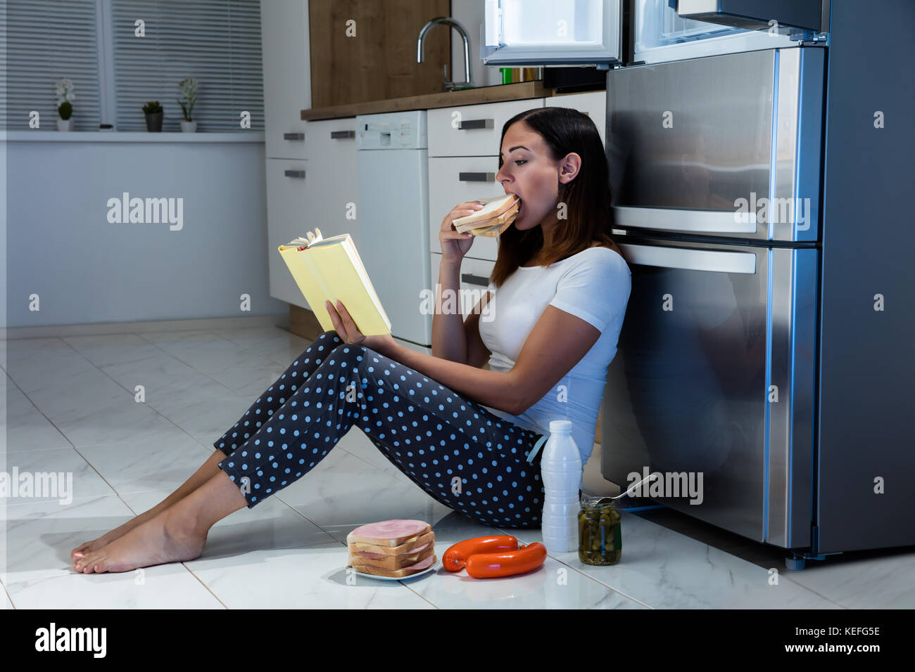 Giovane donna libro lettura mentre mangia sandwich in cucina Foto Stock