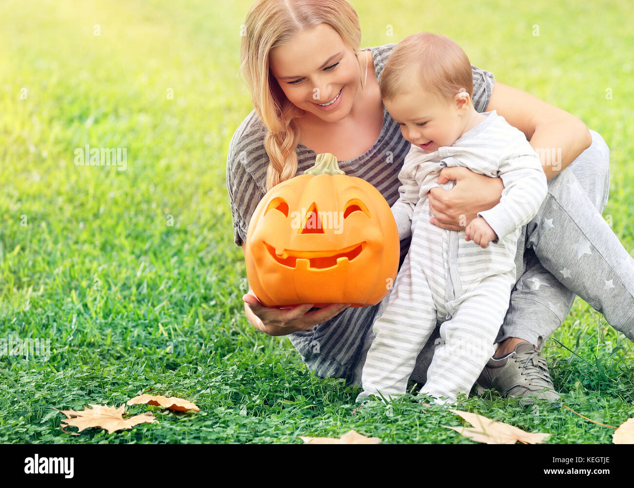 Felice madre con bambino più piccolo all'aperto, seduto sull'erba verde campo e giocando con arancione zucca intagliata con volto sorridente,godendo di halloween h Foto Stock