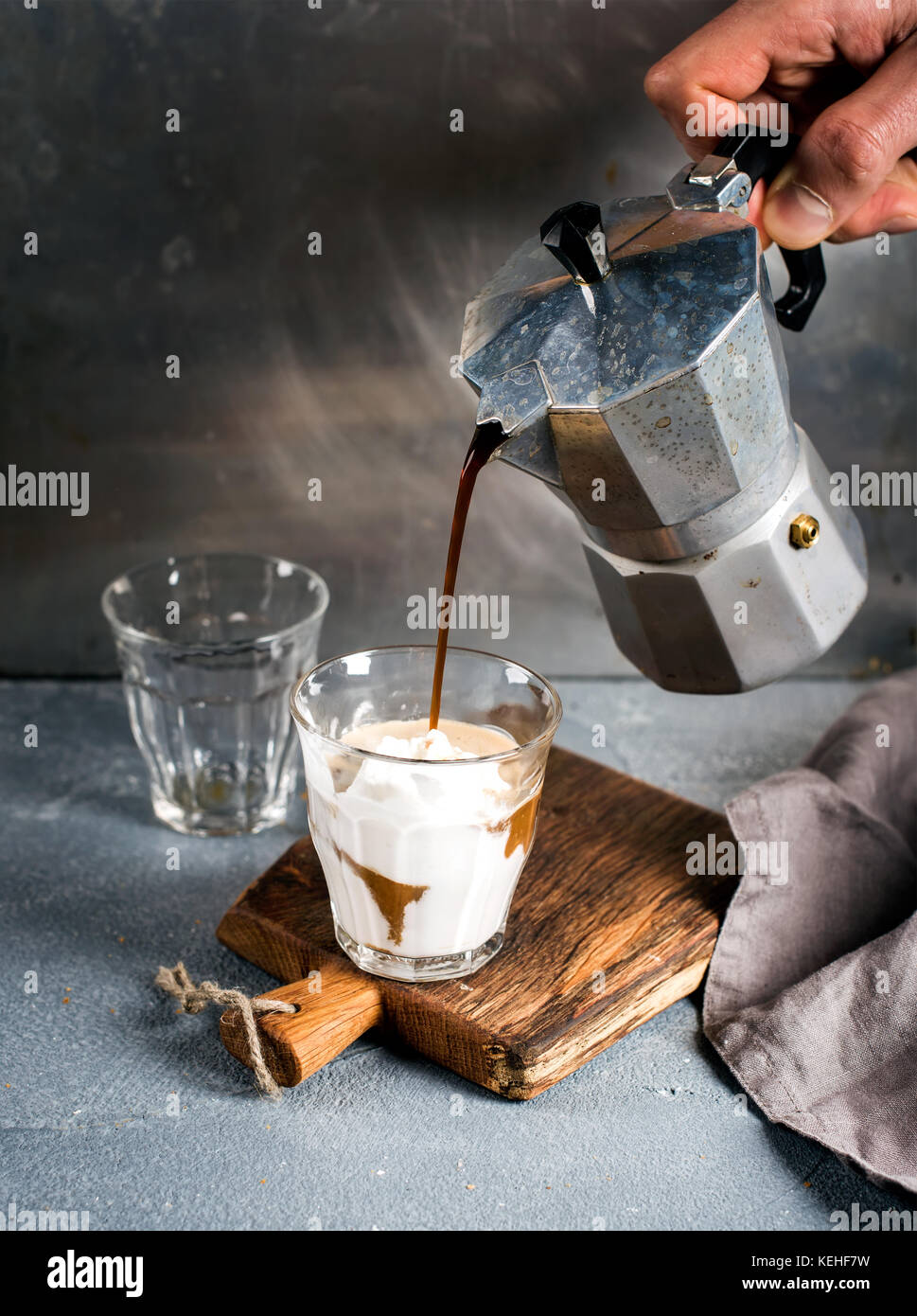 Un bicchiere di caffè con gelato su un rustico di legno. drink è versata da un acciaio moka italiana pot detenute da mano d'uomo, lo sfondo grigio Foto Stock