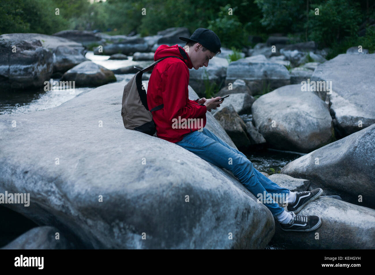 Uomo caucasico seduto sulla roccia in fiume testando sul telefono cellulare Foto Stock