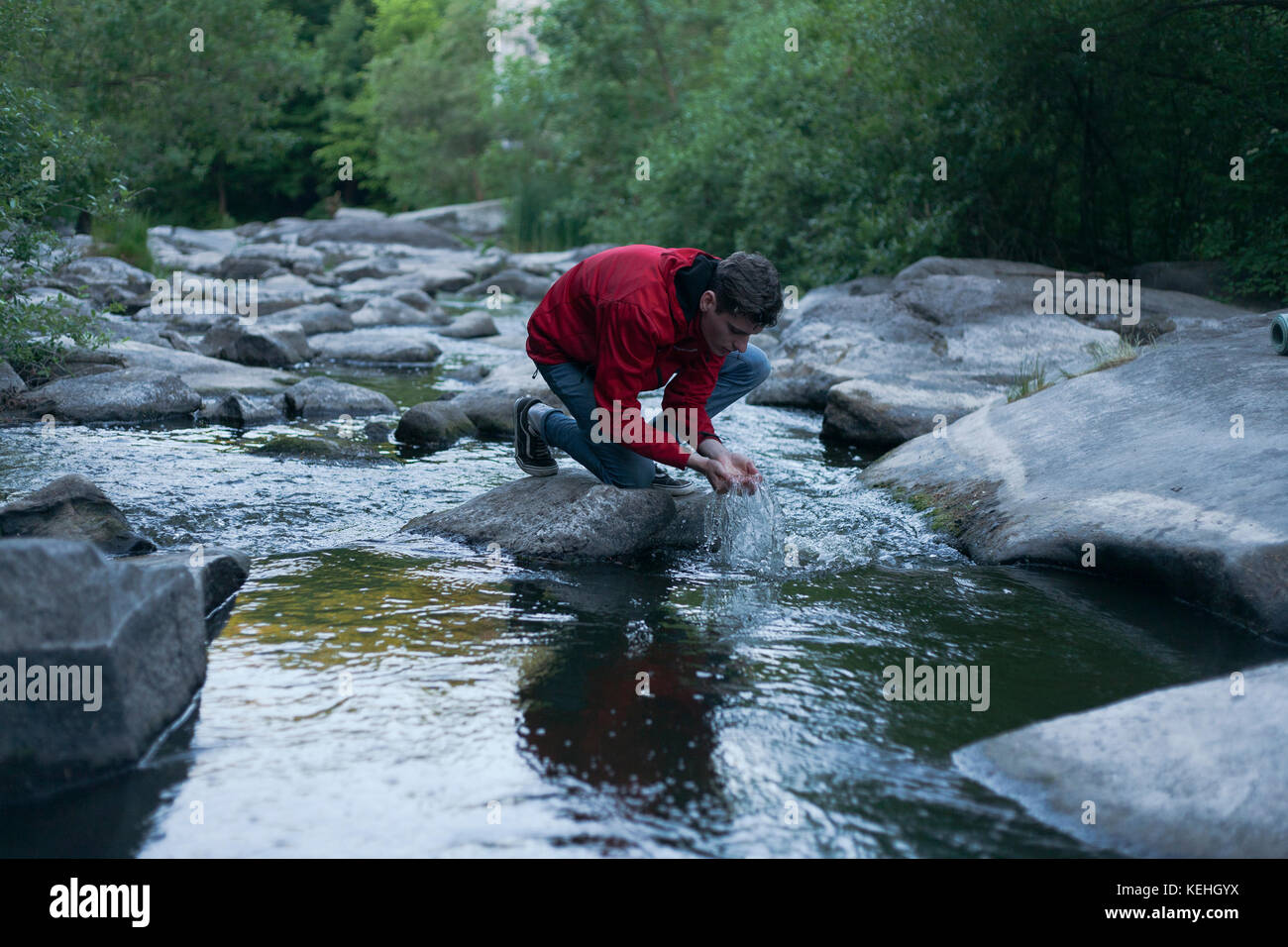 Uomo caucasico inginocchiato sulla roccia nel fiume che cupola l'acqua con le mani Foto Stock