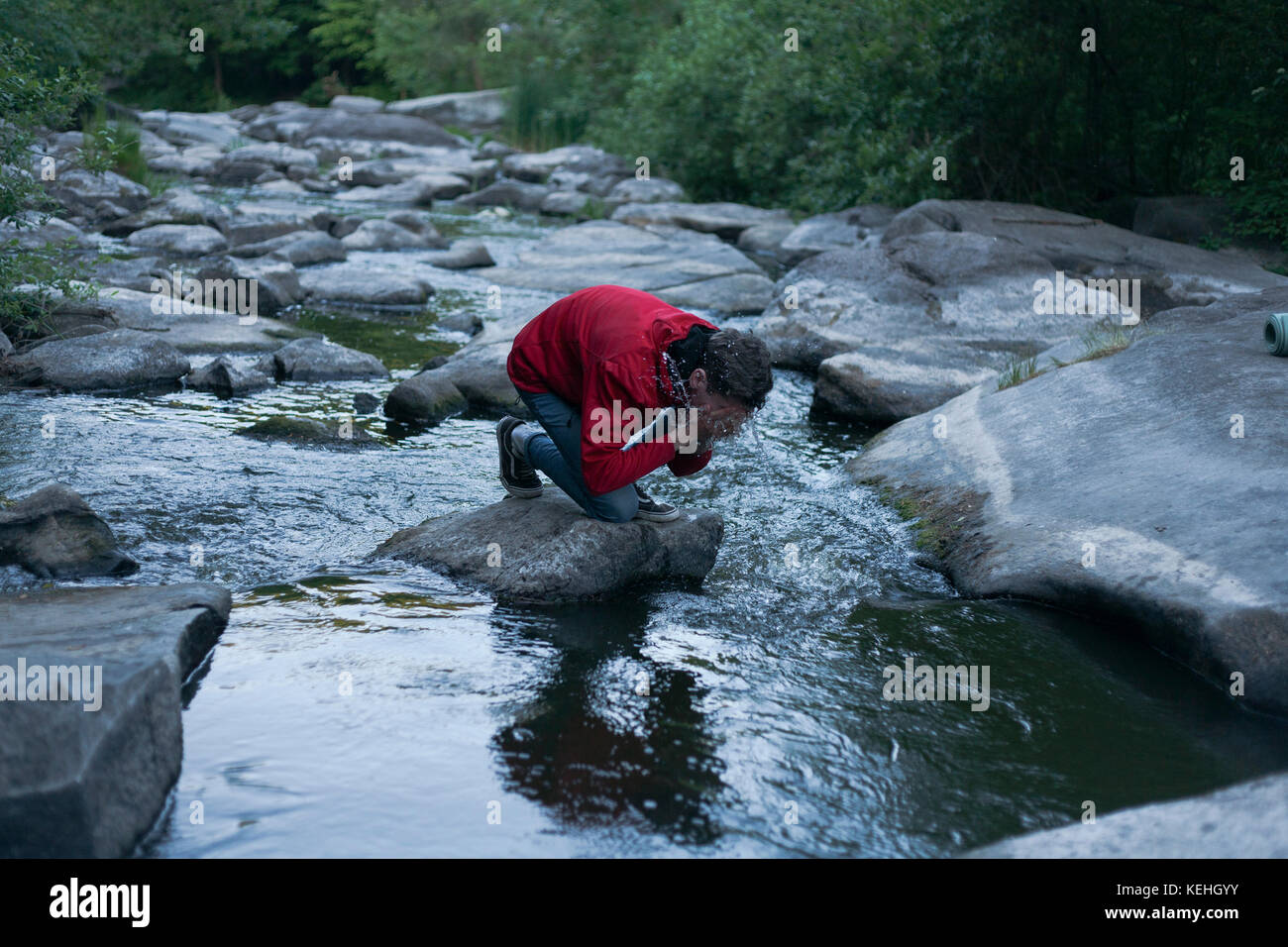 Uomo caucasico inginocchiato sulla roccia nel fiume che spruzzi acqua sulla faccia Foto Stock