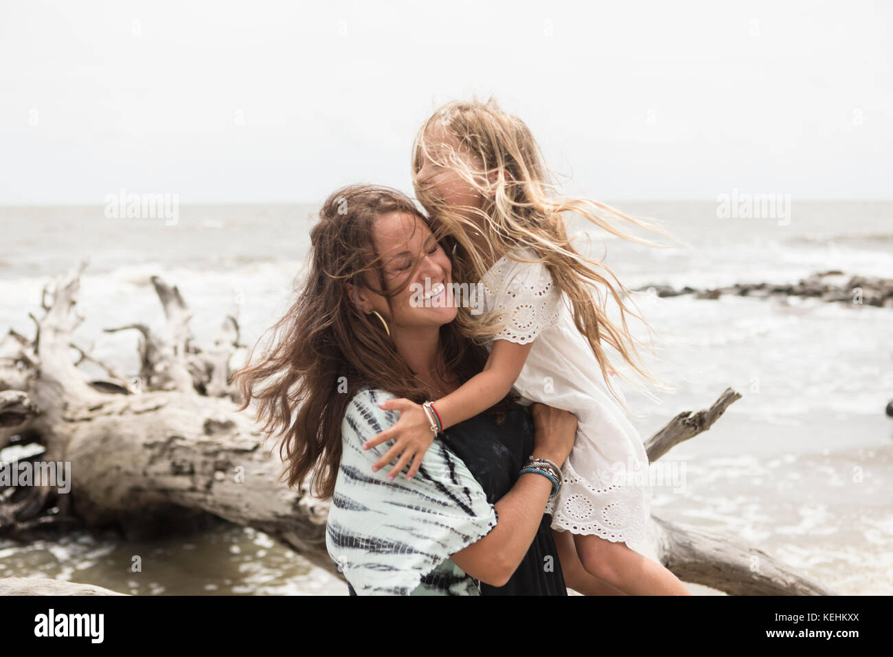 Madre caucasica abbracciando figlia vicino driftwood sulla spiaggia Foto Stock