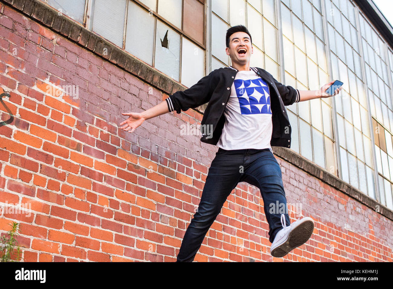 Uomo cinese che salta per gioia vicino muro di mattoni che tiene cellulare Foto Stock