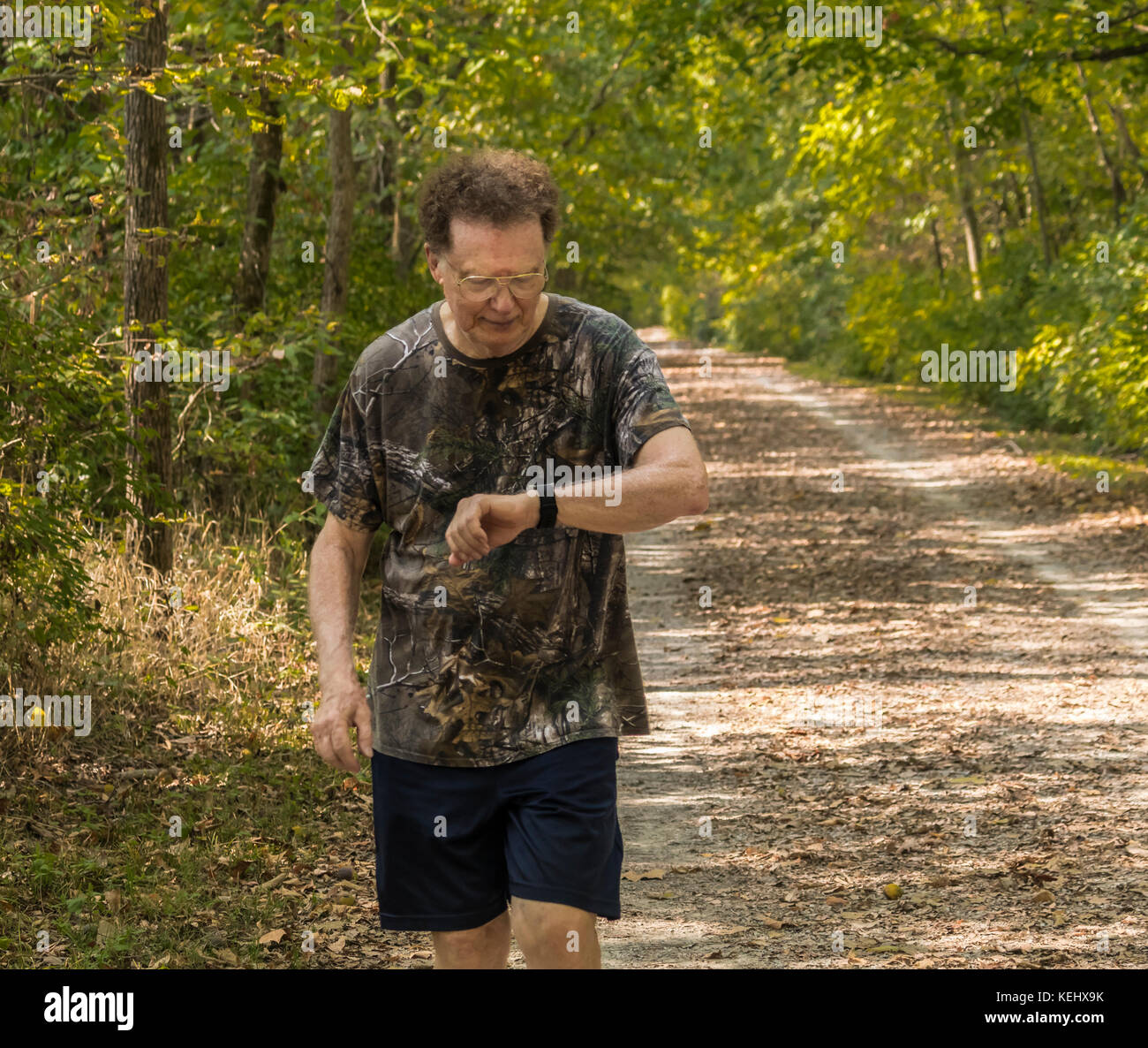 Uomo anziano jogging sul sentiero mentre guardando al suo esercizio bocconcino; gli alberi su entrambi i lati Foto Stock