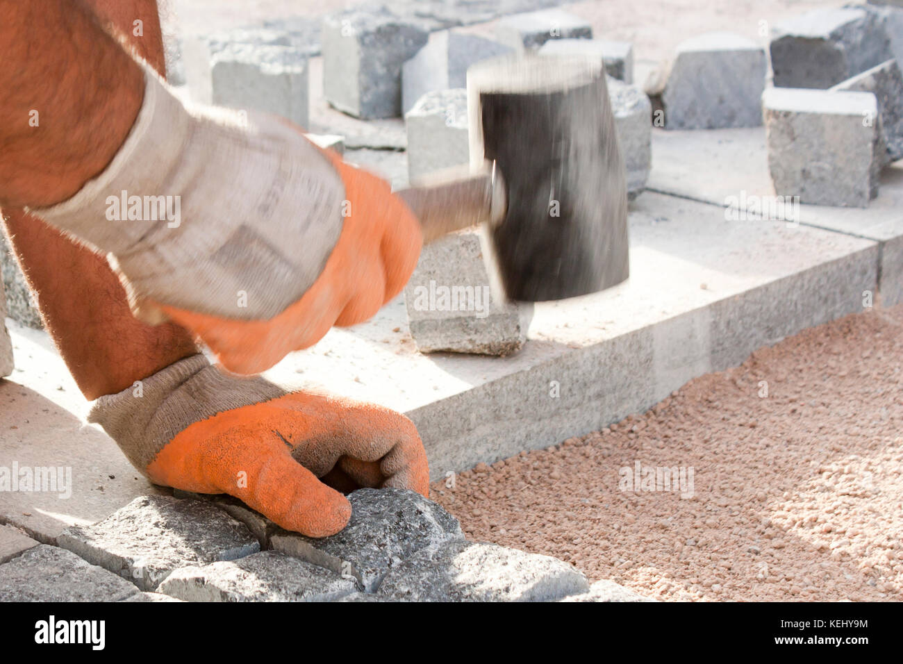Le mani nei guanti di un lavoratore builder posa sui ciottoli di granito imp con un mazzuolo di gomma martello e strada di pavimentazione marciapiede sul sito in costruzione Foto Stock