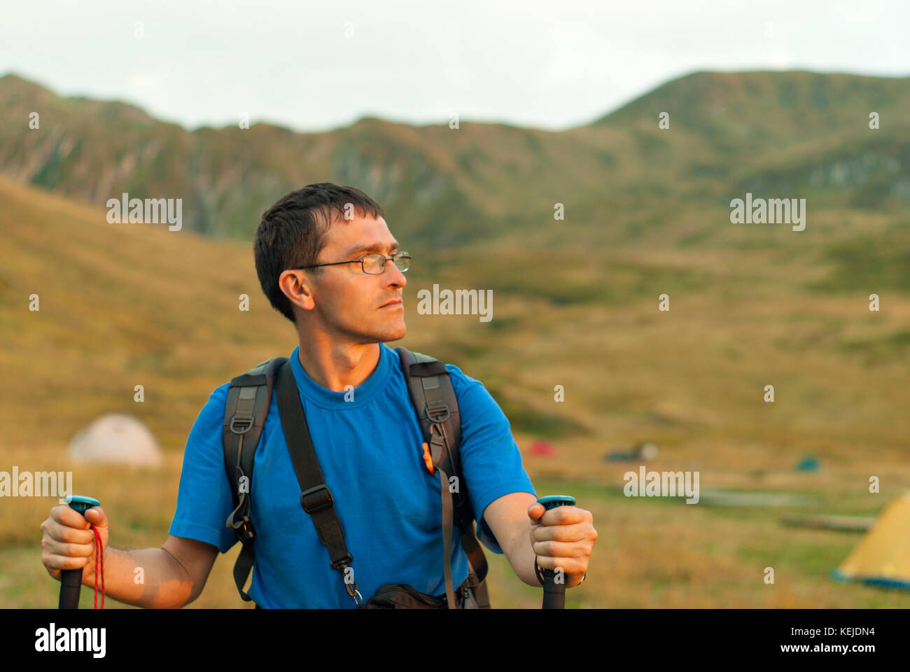 Un uomo con uno zaino, in una maglietta blu e occhiali, con bastoni per il nordic walking è fotografata da nastro contro lo sfondo di una sfocata mounta Foto Stock