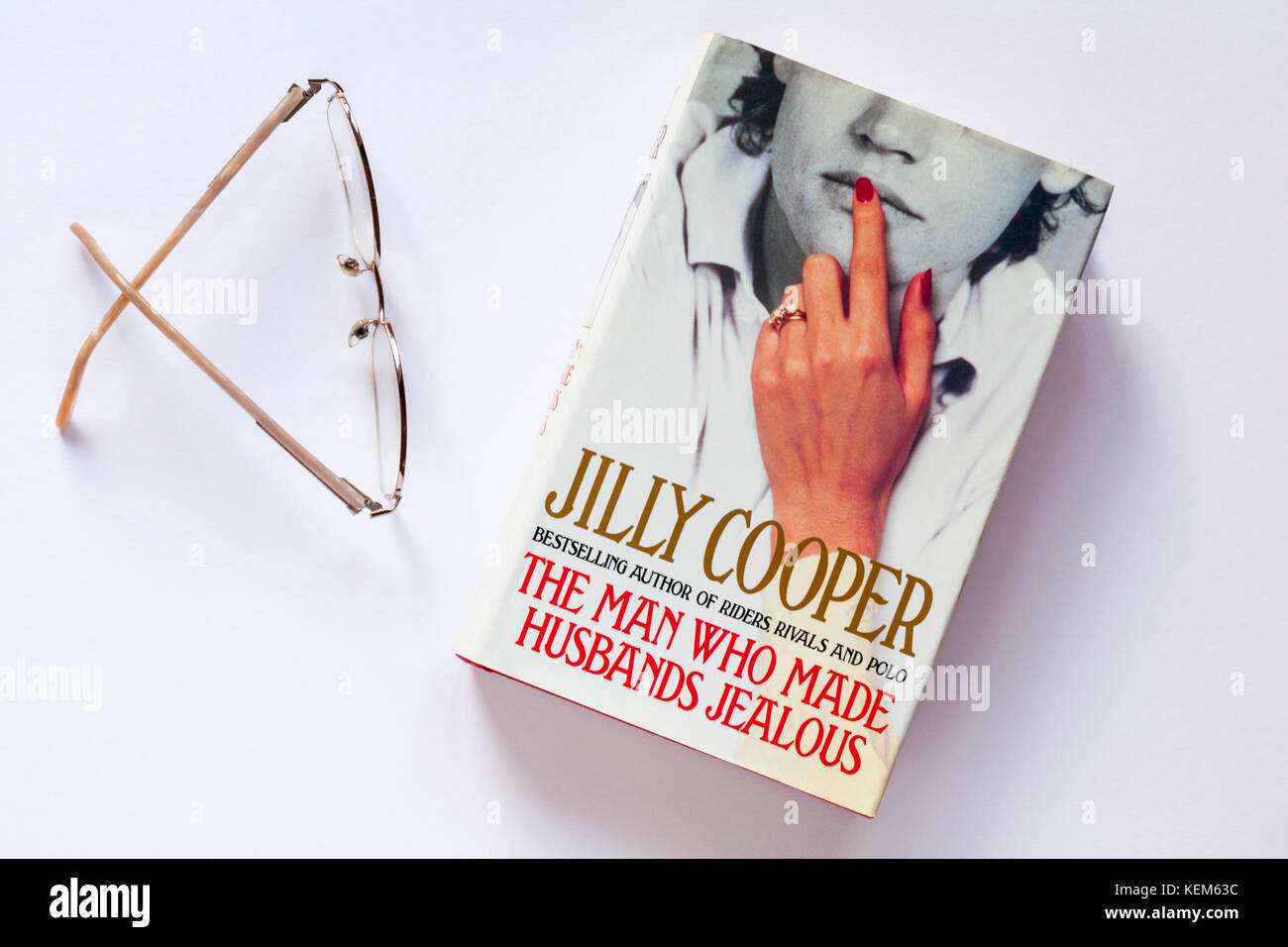 L'uomo che ha fatto i mariti gelosi libro di Jilly Cooper con gli occhiali da lettura isolati su sfondo bianco Foto Stock