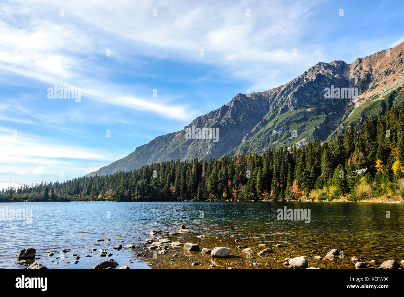 Autunno magnifico paesaggio di montagna con un lago e una foresta ai piedi delle montagne rocciose contro il cielo blu Foto Stock