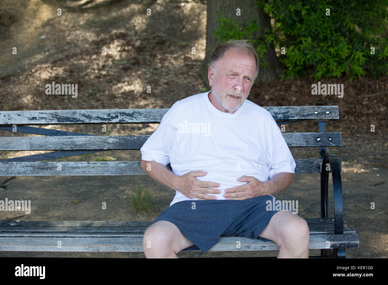 Closeup ritratto, vecchio uomo attanaglia lo stomaco, con dolori di pancia, seduti su una panchina, isolata all'esterno, verdi alberi dello sfondo. Pieno dopo un pasto o il dolore Foto Stock