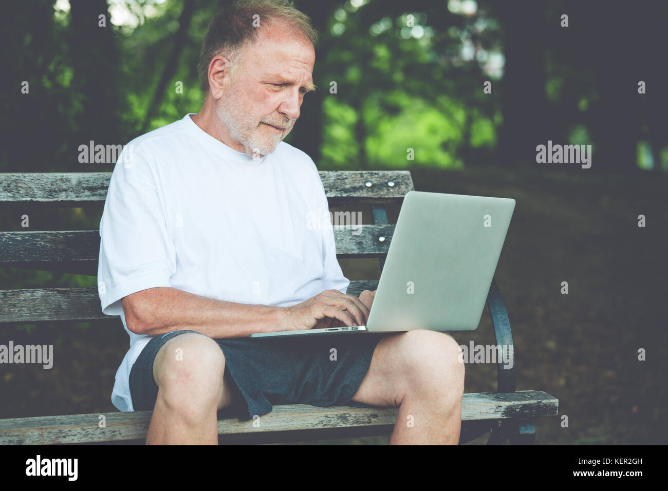 Closeup ritratto, senior uomo maturo in maglia bianca a scrivere, navigazione digitale computer laptop, sfondo isolato di sunny outdoor, verdi alberi na Foto Stock