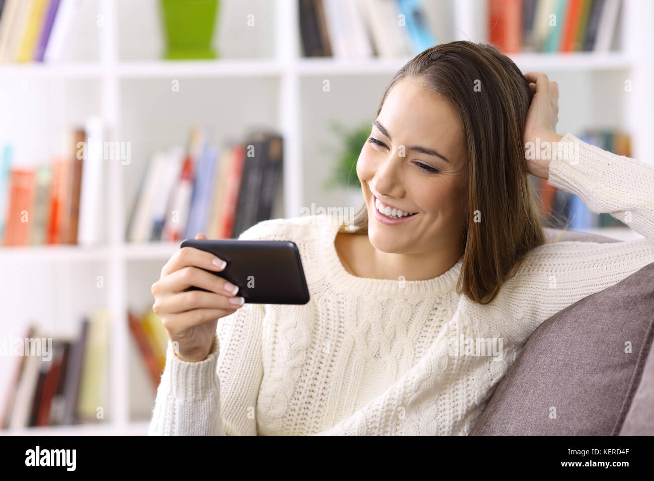 Felice ragazza guardare contenuti in streaming su linea in uno smart phone seduto su un divano in inverno a casa Foto Stock