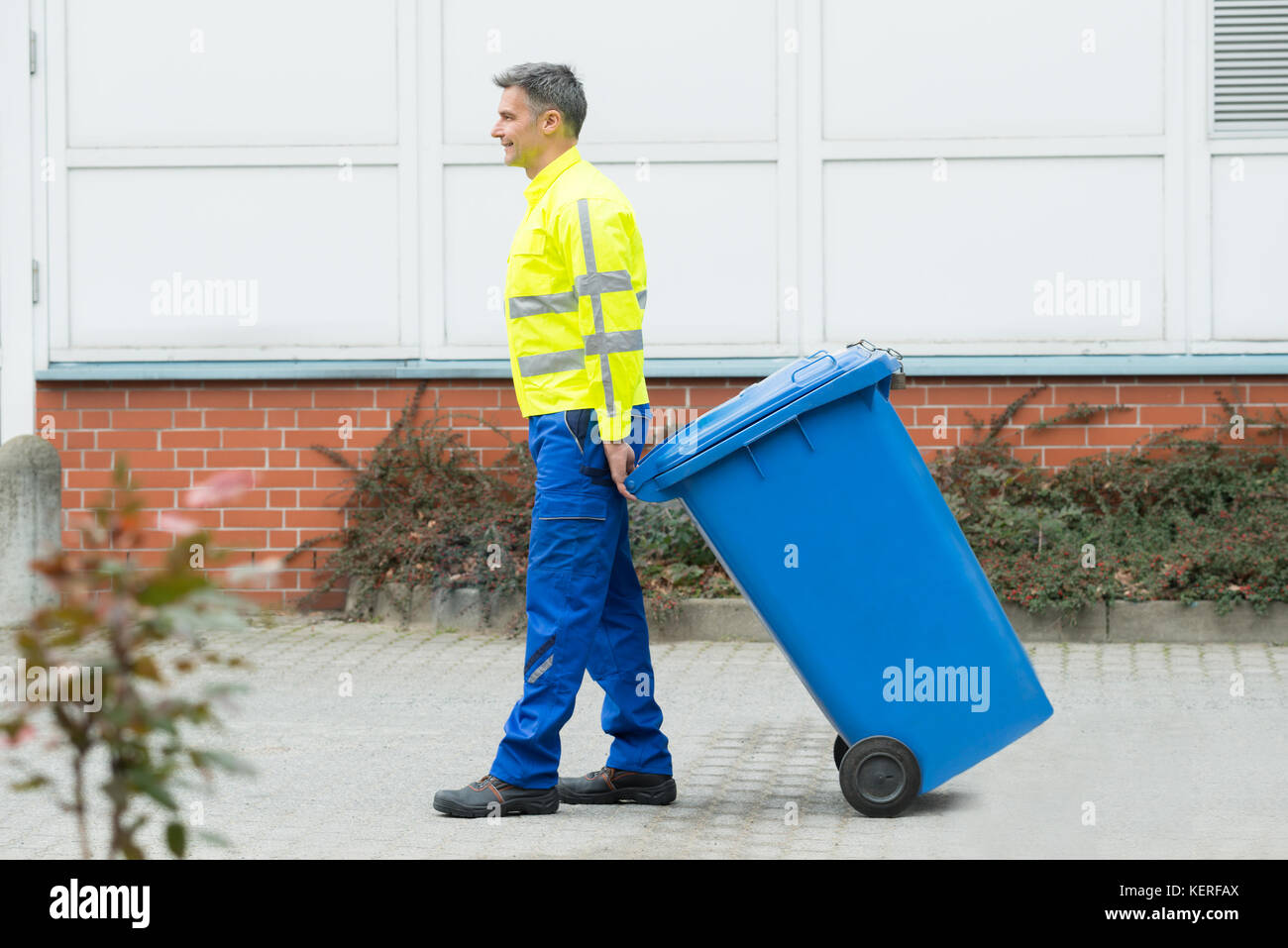 Felice lavoratore maschile camminare con il bidone della spazzatura in strada durante il giorno Foto Stock