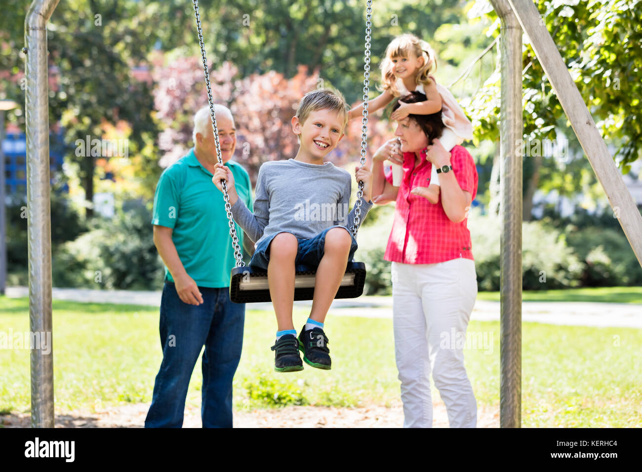 Capretto felice avendo divertimento su Swing con i nonni in piedi dietro nel Parco Foto Stock