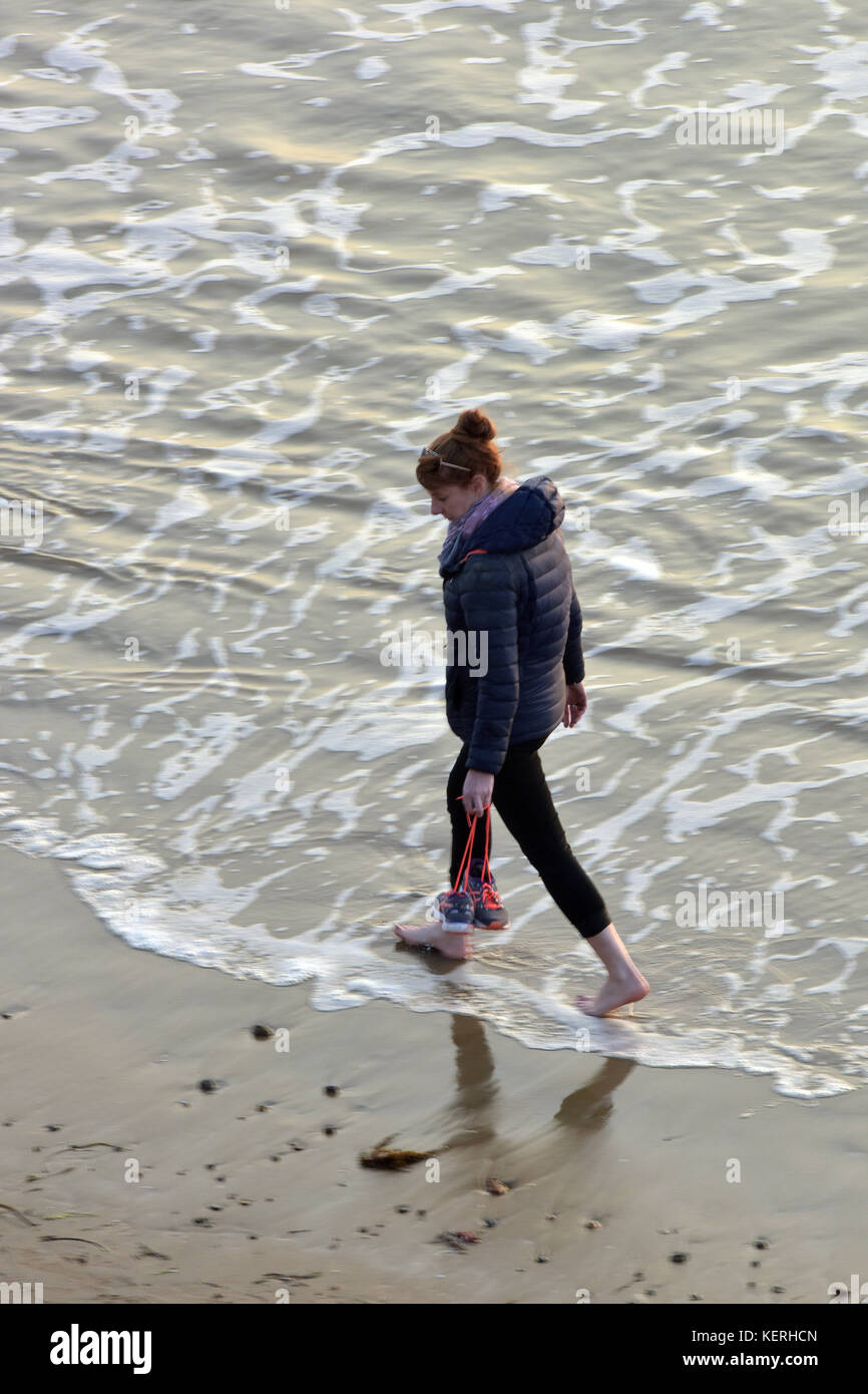 Una giovane donna che cammina lungo su di una spiaggia di sabbia in acqua pagaiando portando le sue scarpe per evitare loro bagnarsi nel mare ad alta marea. pala in mare Foto Stock