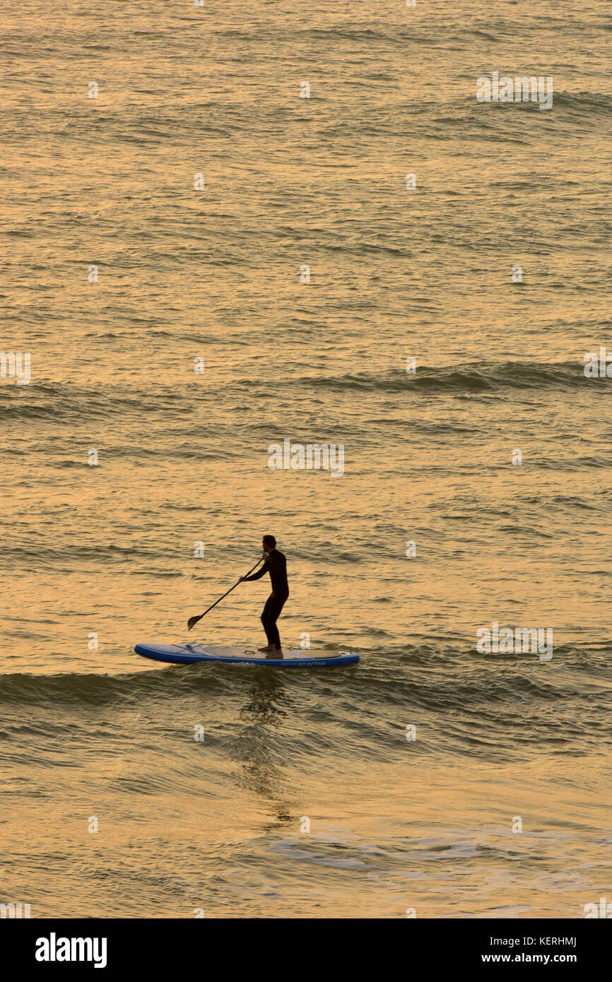 Un uomo con una pagaia o scheda di paddle boarder tra le onde e il surf al tramonto tenendo una racchetta e pagaiando sopra le onde e demolitori. Foto Stock