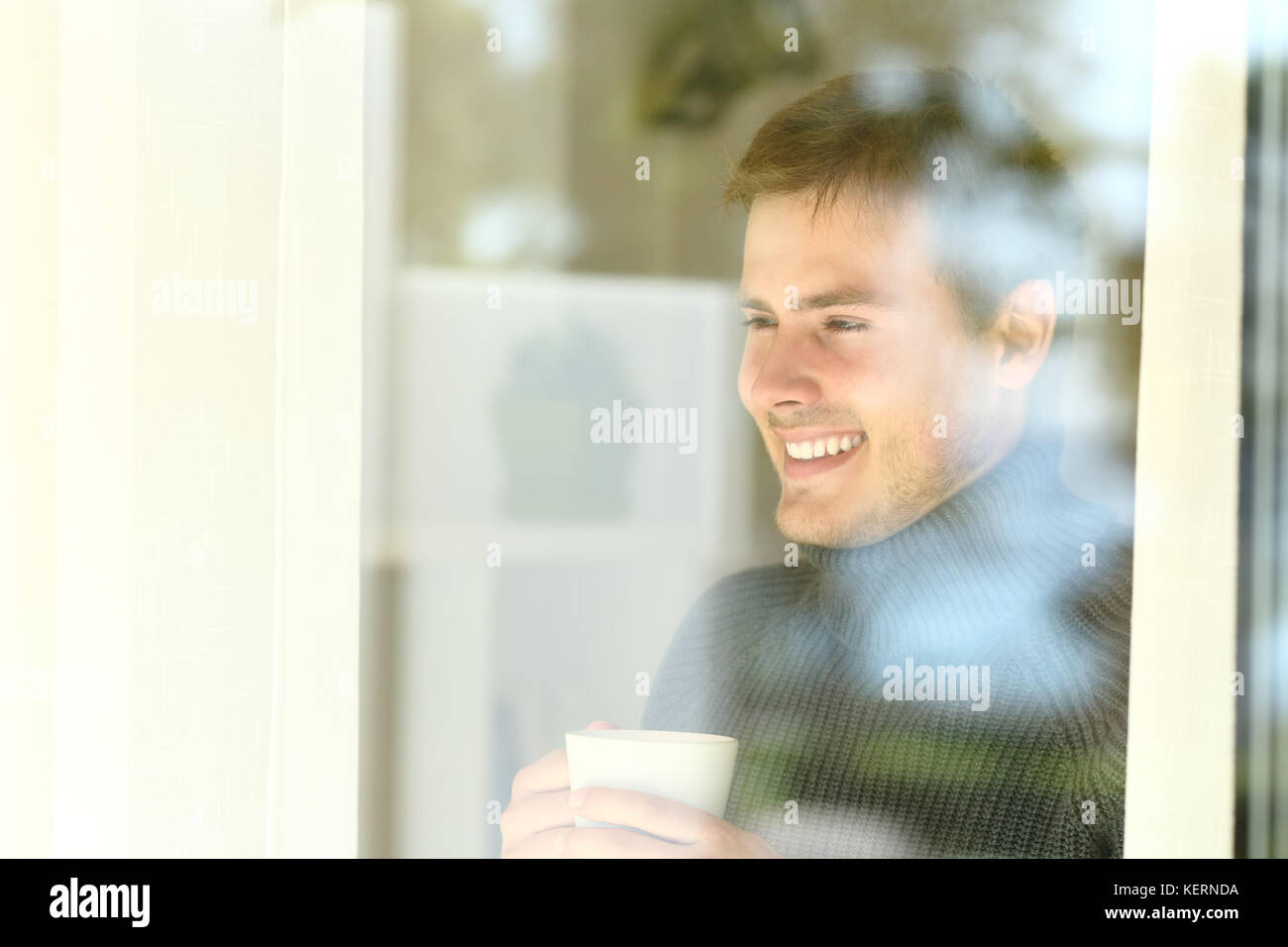 Uomo felice tiene una tazza di caffè guardando attraverso una finestra in piedi a casa o in albergo in inverno Foto Stock