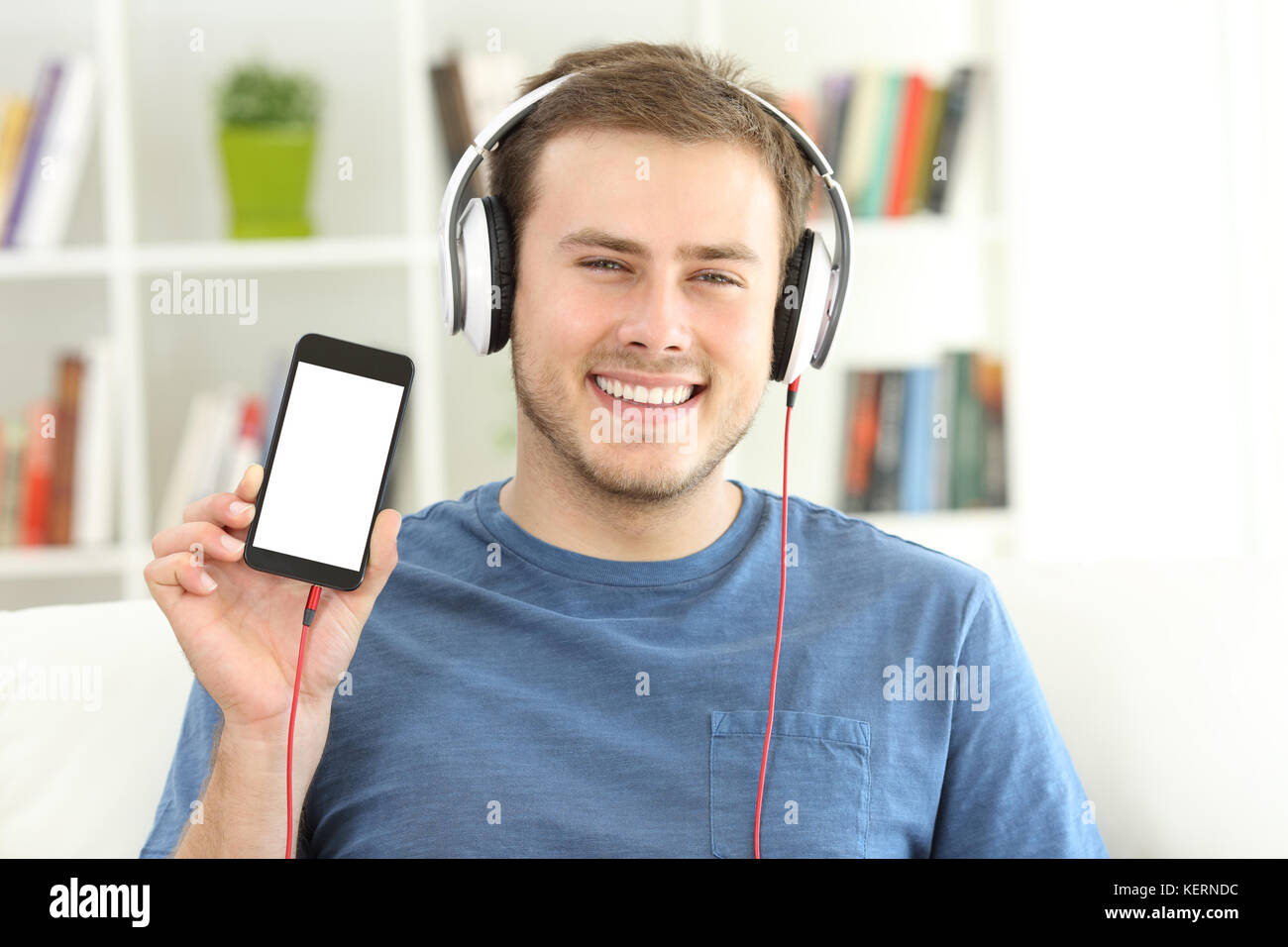 Vista frontale - Ritratto di un uomo felice l'ascolto di musica e che mostra un fustellato smart phone schermo seduto su un divano a casa Foto Stock