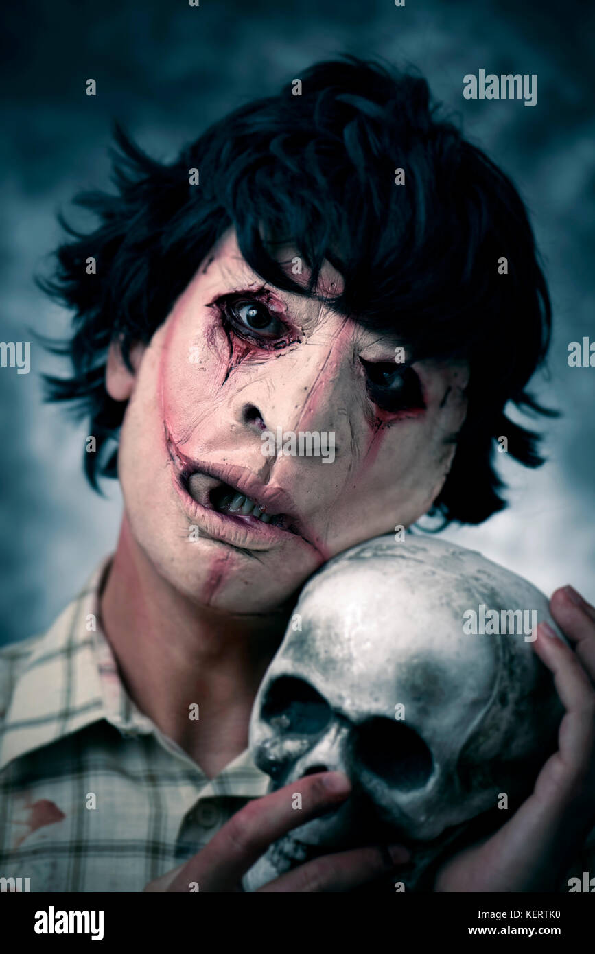 Primo piano di un pauroso sfigurato uomo che indossa un frastagliato e maglietta sporca con macchie di sangue, tenendo un cranio nelle sue mani Foto Stock