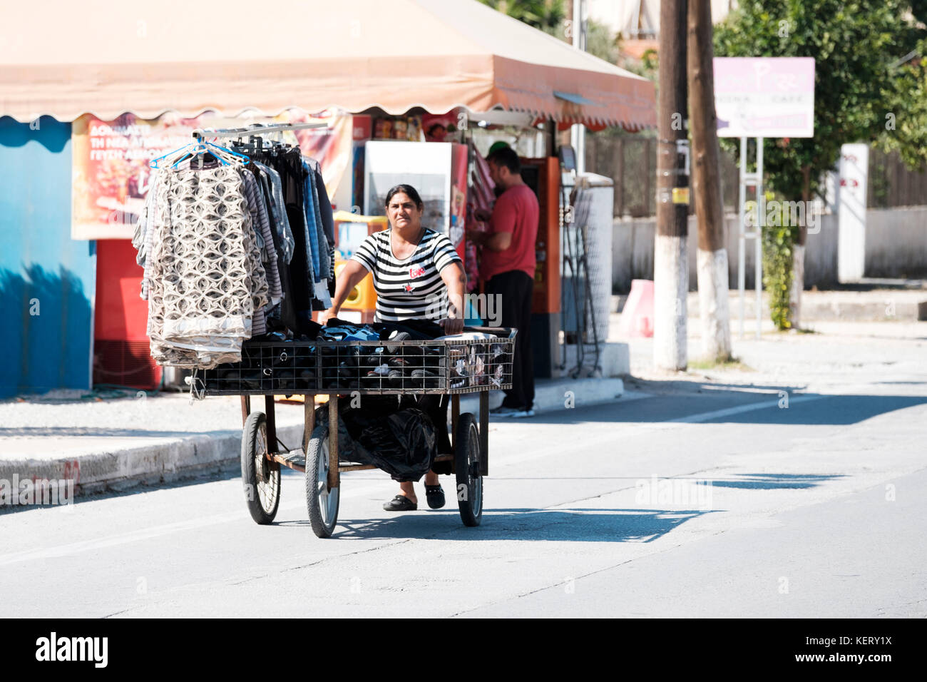 Una donna greca anziana che spinge un carrello a tre ruote lungo la strada principale. hte cart è caricato con gli articoli di abbigliamento che sta cercando di vendere Foto Stock
