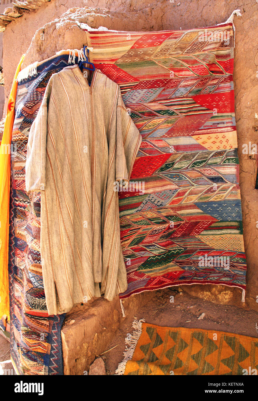 Tradizionale uomini marocchini djellaba abbigliamento e tappeto di lana sulla parete di argilla, souk in ait-ben-haddou kasbah, Marocco, Africa del nord Foto Stock