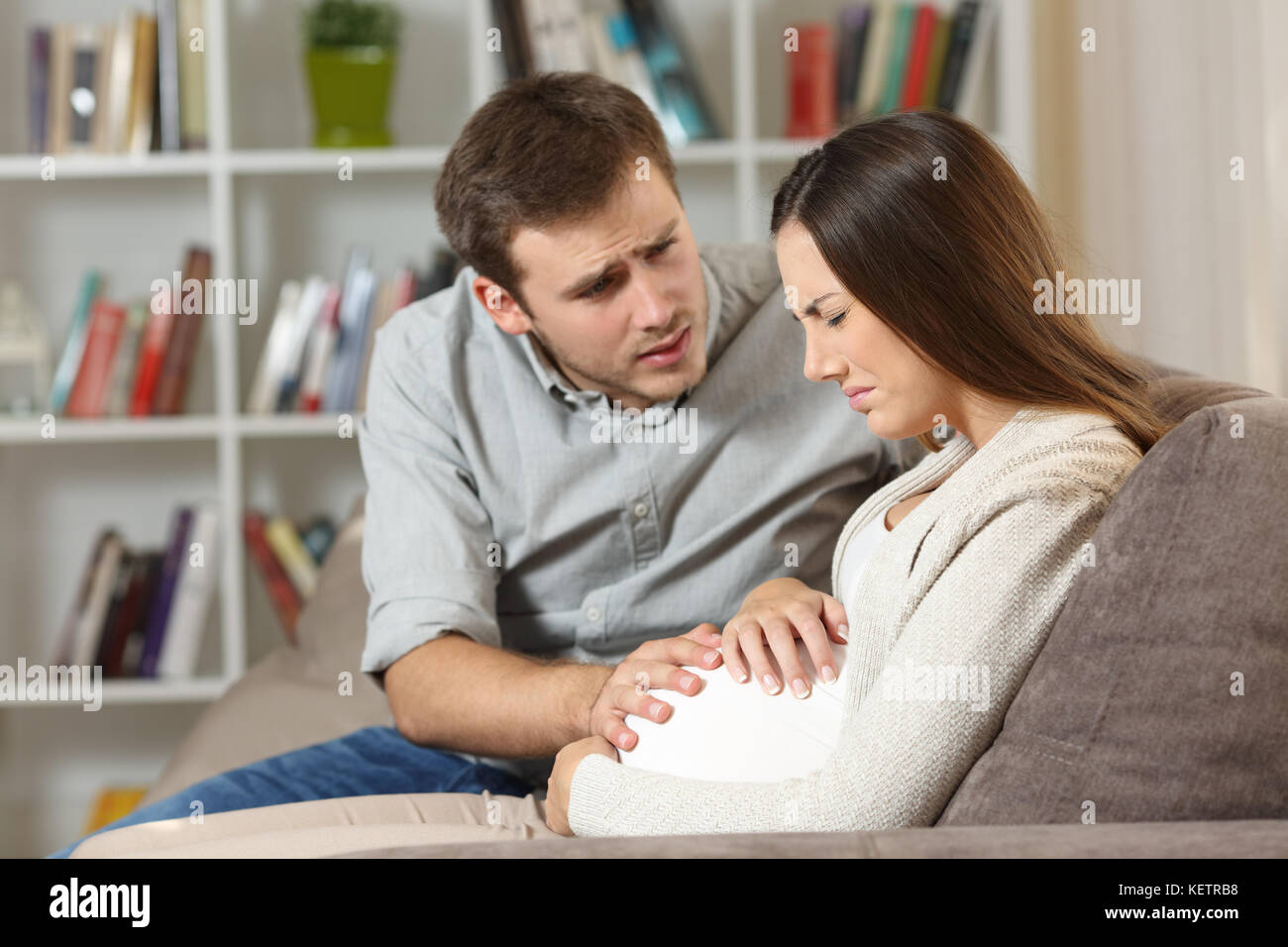 Donna incinta soffre il mal di pancia e il marito consolante la sua seduto su un divano a casa Foto Stock