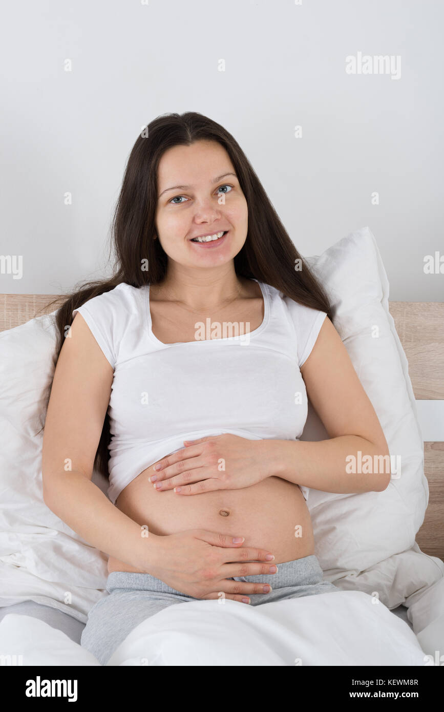 Felice donna incinta seduta sul letto di casa Foto Stock