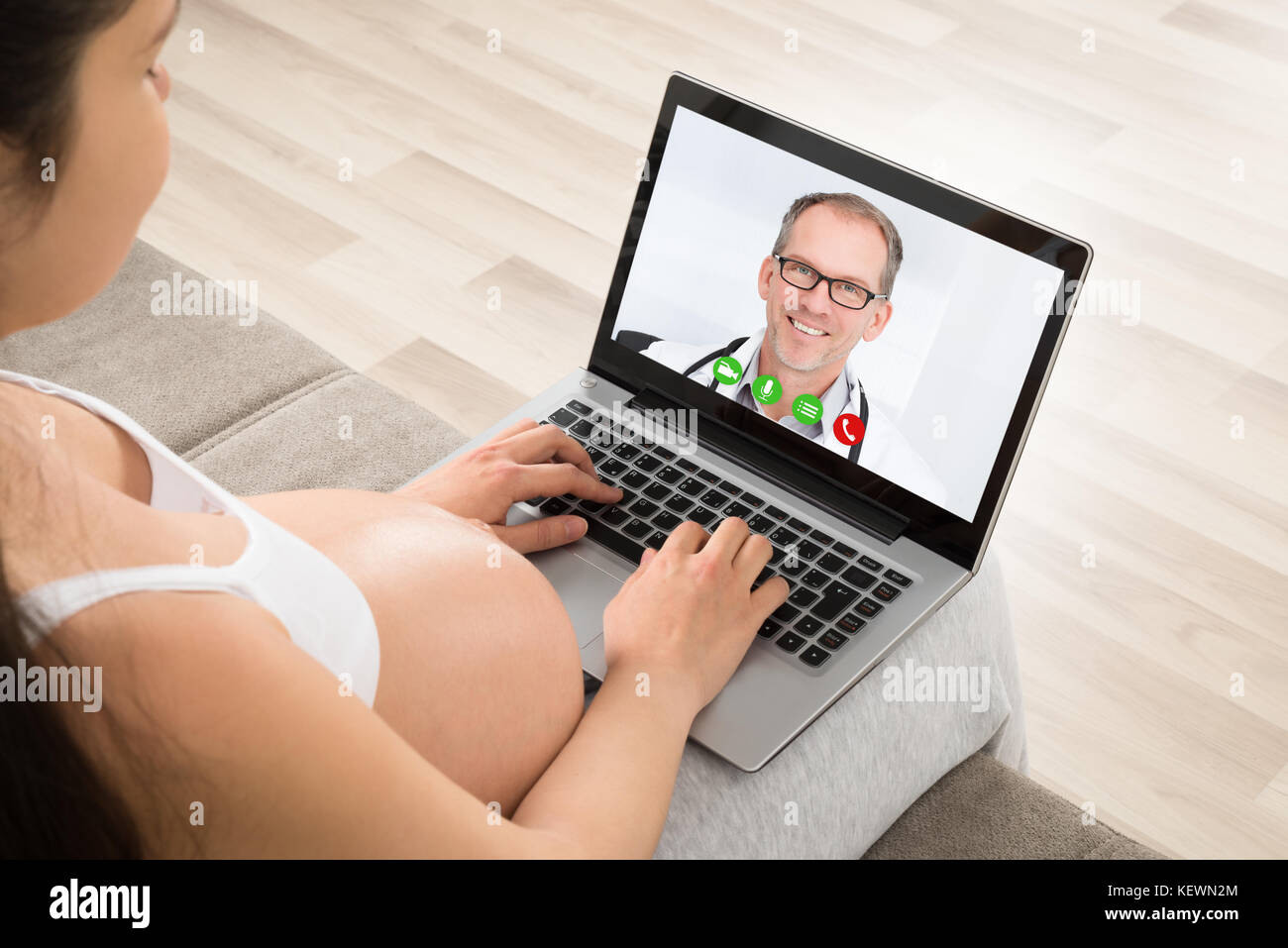 Donna incinta seduta sul divano la videoconferenza con medico sul portatile a casa Foto Stock